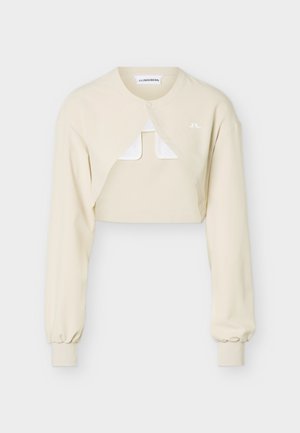 Μπεζ κοντό μπολερό με μακριά μανίκια, εμπρόσθιο κούμπωμα με κλιπ, ριμπ μανσέτες και μικρό λευκό λογότυπο στο αριστερό μέρος του στήθους.