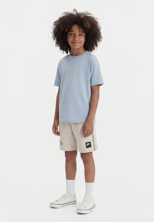 Barn med krøllet hår iført en lyseblå t-shirt, beige Nike Air-shorts, hvide sokker og hvide sneakers, stående og smilende.