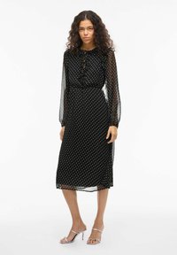 Robe midi à pois noirs avec des manches longues transparentes, un col à volants et une taille cintrée. Texture lisse ; éclat subtil. Talons argentés à lanières.