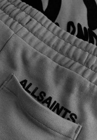 Los pantalones deportivos grises cuentan con una cintura elástica y un pequeño bolsillo. "ALLSAINTS" está bordado en negro en la parte trasera, cerca del bolsillo.