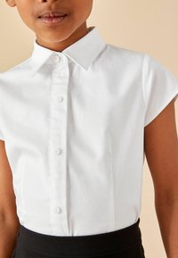Chemise blanche à col avec des manches courtes, dotée d'un devant à boutons et d'une texture lisse, accompagnée d'une ceinture noire en dessous.
