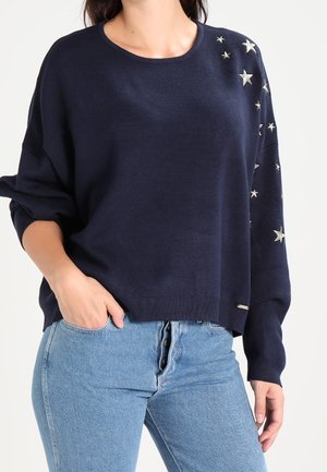 Kobieta ubrana w granatowy sweter z srebrnymi wzorami gwiazdek na jednym ramieniu oraz jasnoniebieskie jeansy z wysokim stanem, jedna ręka w kieszeni.