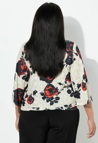 Květinová blůza s krémovým pozadím a vzorem červených a černých růží, s tříčtvrtečními zvonovými rukávy a volným střihem. Pod ní jsou nošeny černé kalhoty.