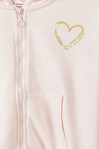 Sudadera con cremallera de un suave tejido en color rosa claro, que presenta un corazón bordado en dorado y blanco con el texto "Siempre Encantadora". Incluye bolsillos frontales.