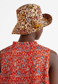 Chapeau seau à motif floral avec un fond brun foncé, orné de marguerites blanches et jaunes. Le modèle porte un haut floral coloré et texturé.