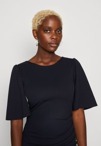 Jūras zila kleita ar īsām, izplestām piedurknēm, apaļu dekoltē un savilkuma detaļām jostas vietā. Gluda auduma ar pieguļošu siluetu.