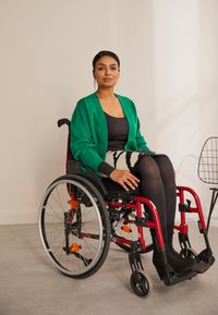 Rode rolstoel met grijze wielen en oranje accenten. Individu draagt een groene cardigan over een zwarte top en een gestreepte rok met zwarte panty.