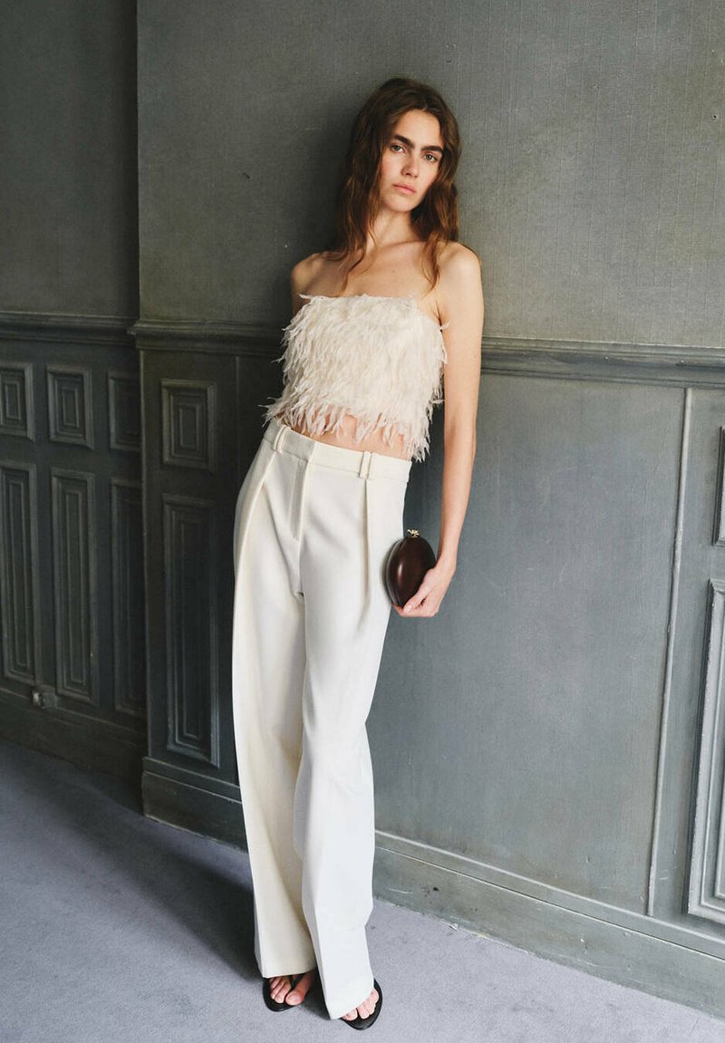 Femme portant un top bustier blanc orné de plumes, un pantalon large taille haute, des sandales noires, tenant une petite pochette sombre, appuyée contre un mur gris.