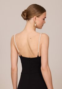 Femme aux cheveux bruns en chignon portant une robe noire avec des bretelles en perles et de grandes boucles d'oreilles pendantes en perles, vue de dos sur un fond beige.