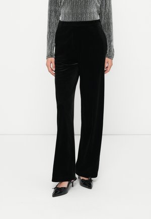 Femme portant un pantalon large en velours noir, un top argenté à manches longues et des escarpins noirs à bouts pointus avec des nœuds.