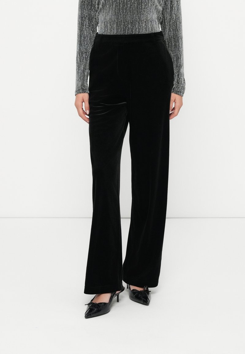 Femme portant un pantalon large en velours noir, un top argenté à manches longues et des escarpins noirs à bouts pointus avec des nœuds.