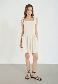 White, sleeveless dress with a square neckline, tie-shoulder straps, and gathered skirt. Paired with black strappy sandals.
