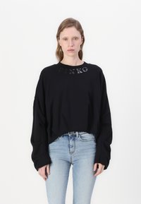 Felpa nera oversize con logo "PINKO" in materiale lucido lungo il colletto, abbinata a jeans aderenti blu chiaro.