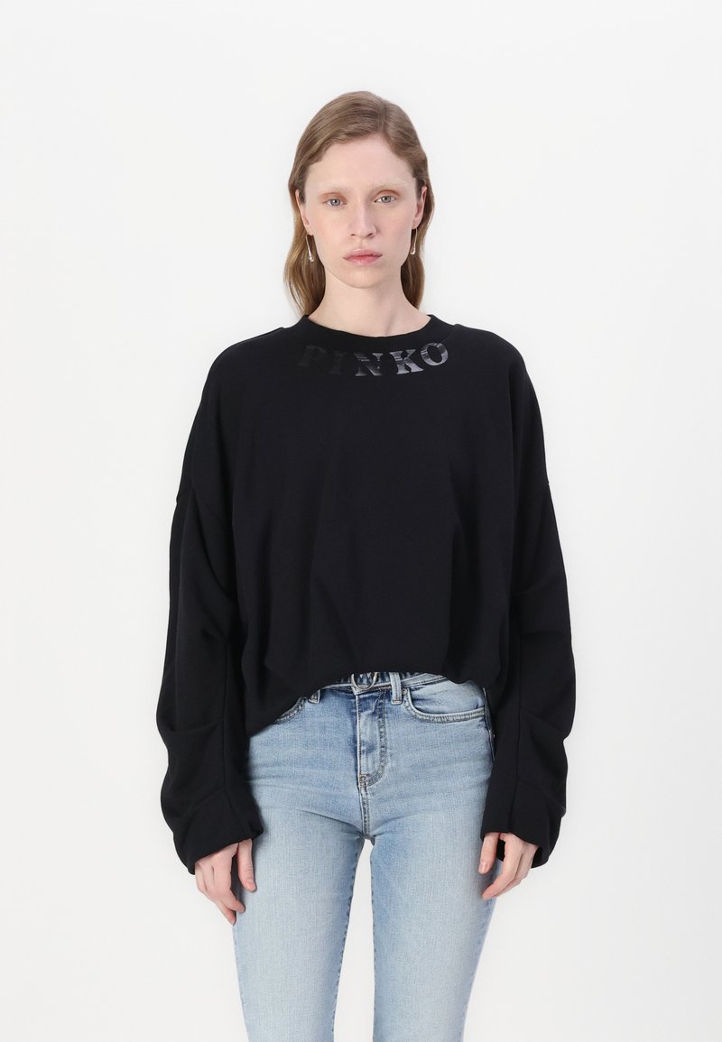 Felpa nera oversize con logo "PINKO" in materiale lucido lungo il colletto, abbinata a jeans aderenti blu chiaro.
