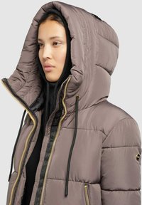 Pufferjack in licht taupe met een grote, gequiltte capuchon, zwarte trekkoorden, gouden rits en geribbelde kraag, met een logo-accent op de mouw.
