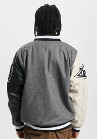 Grå ullkropp jakke med svarte skinn ermer og kremfargede detaljer. Har en stripete krage og broderte lapper på ermene.