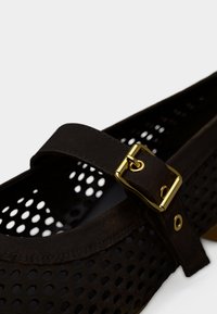 Chaussures marron foncé perforées avec une boucle dorée et une bride en cuir. Le design est arrondi avec un intérieur lisse.