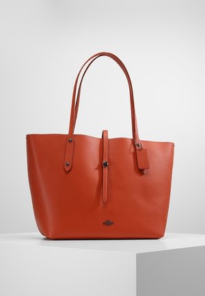 Stor orange lædertaske med to skulderremme, en central knaprem og en lille hængende mærkat på et hvidt podium.