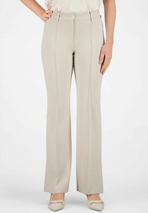 Vrouw draagt beige hoge taille, wijde geruite broek met voorsluiting en knopen, gecombineerd met beige spitssluitende hakken met strikjes.