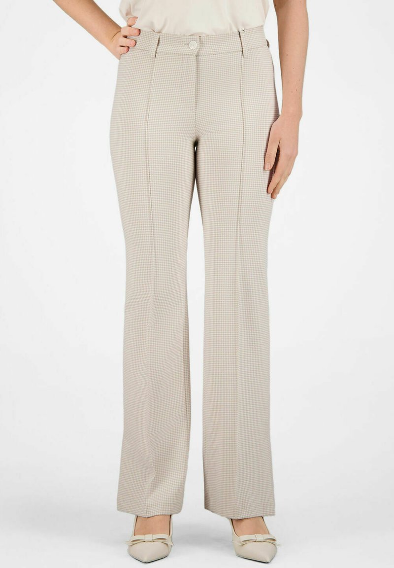 Vrouw draagt beige hoge taille, wijde geruite broek met voorsluiting en knopen, gecombineerd met beige spitssluitende hakken met strikjes.