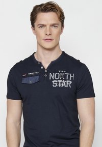 Tamno plava majica s kratkim rukavima i henley izrezom, sadrži džep od trapera, tiskani logo "NORTH STAR" i kontrastne naglaske na ramenima.