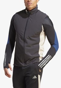 Chaqueta deportiva híbrida en negro, beige y azul. Cuenta con un cuello alto, cierre con cremallera y paneles texturizados con franjas reflectantes.