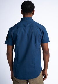 Korte mouwen knoopsluiting overhemd van donkerblauwe katoen. Heeft een puntige kraag, achterpand yoke en een zwarte patch op de linker schouder.