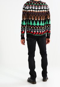 Homme portant un pull de Noël coloré orné de visages du Père Noël, de rennes, de cannes en bonbon, d'arbres, de chaussettes et de guirlandes lumineuses, associé à un jean noir et des chaussures.