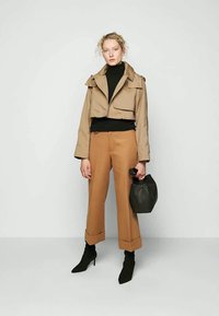 Cropped beige jacket with large lapels, black turtleneck, tapered light brown trousers, and black pointed ankle boots, holding a black bag.