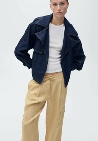 Donker jeansjack met oversized kraag, voorzien van een boxy pasvorm en contrasterende stiksels. Gecombineerd met lichtbeige wijdere broek.