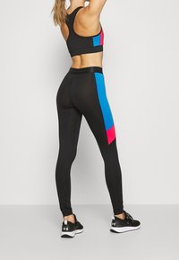 Černé sportovní legíny s růžovými a modrými bočními panely, kombinované s černým sportovním podprsenkovým topem. Tkanina vypadá hladce a přiléhavě. Černé tenisky.