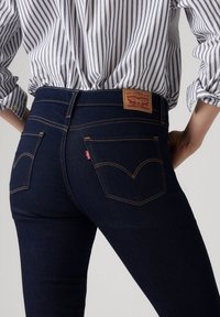 Vaqueros de mezclilla azul oscuro con cintura alta, costuras visibles y dos bolsillos traseros. Una etiqueta marrón con el logo aparece en la cintura.
