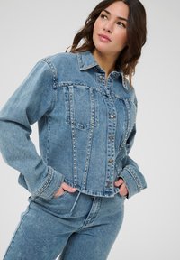 Chaqueta de mezclilla en azul claro, con remaches plateados en las costuras, un cuello puntiagudo y cierre de botones en la parte delantera. Manga larga y dos bolsillos en el pecho.