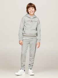 Felpa grigia con logo, abbinata a pantaloni della tuta grigi coordinati. Sneakers bianche con dettagli blu navy. Tessuto morbido, vestibilità rilassata.