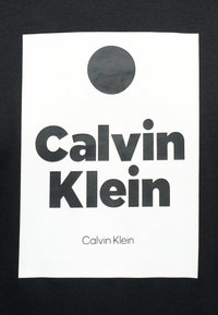 Μαύρος κύκλος πάνω από παχύ έντονο μαύρο κείμενο "Calvin Klein" σε λευκό ορθογώνιο φόντο, τοποθετημένο στο κέντρο μαύρου υφάσματος.