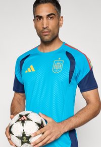 Herrarnas sporttröja i klarblått och marinblått, med ett texturerat mönster och Spaniens emblem, hålls över en vit fotboll med svarta detaljer.