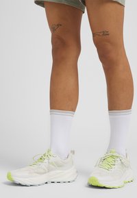 White athletic shoes with light green accents and mesh details, paired with white socks featuring gray trim. Visible leg tattoos on knees.