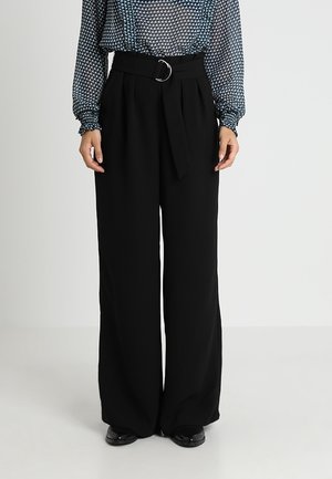 Frau trägt hoch taillierte, weit geschnittene schwarze Hose mit Gürtel und silberner Ring-Schnalle, kombiniert mit einer gemusterten Bluse mit langen Ärmeln.