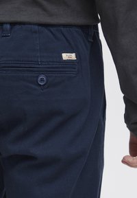 Pantalon bleu marine au toucher lisse, avec une poche arrière, une fermeture à bouton et une petite étiquette de marque sur la ceinture.