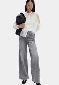 Blusa di pizzo bianco con scollo a volant, abbinata a jeans grigi a vita alta e gamba larga e scarpe con tacco nere. Borsa nera è tenuta in una mano.