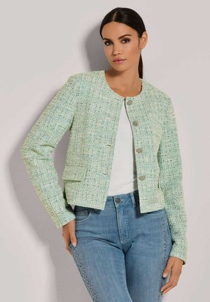 Muntgroene tweedjas met een ronde halslijn, voorzakken en decoratieve zilveren knopen, gecombineerd met lichtblauwe jeans met studs.