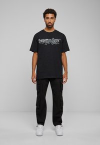 Črna bombažna majica s grafičnim dizajnom kuščarja, kombinirana s črnimi cargo hlačami in belimi supergami. Model stoji pred sivo ozadjem.