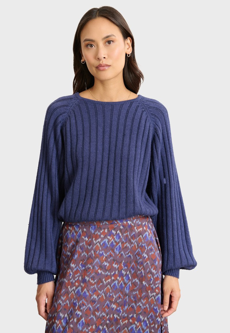 Frau in einem gestreiften navyblauen Pullover mit Ballonärmeln und einem gemusterten Rock in Lila-, Rost- und Weißtönen vor einem hellen Hintergrund.