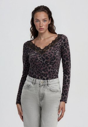 Giovane donna con lunghi capelli scuri che indossa un top a maniche lunghe con stampa leopardata, scollo in pizzo nero e jeans grigio chiaro a vita alta.