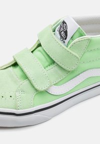 Zapatillas de ante verde y lona con dos correas de Velcro, detalles en blanco y ribete negro. Presenta una superficie texturizada y un cuello acolchado.