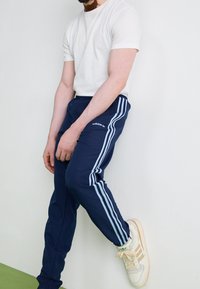 Mörkblå träningsbyxor med ljusblå ränder på sidorna, i kombination med en enkel vit t-shirt och beiga sneakers. Slät tygtextur.