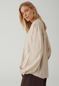 Long-sleeve, beige blouse made of a soft fabric. Features a relaxed fit, gathered shoulders, and subtle side pleats at the back.