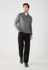 Un sweat à zip gris avec des poignets côtelés, associé à un pantalon noir et des chaussures de sport foncées. Comprend un petit logo jaune sur la poitrine.