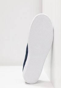 Baskets bleu marine avec dessus en textile texturé, semelle en caoutchouc blanche présentant un motif de bande de roulement en zigzag, placées en angle sur un fond blanc.