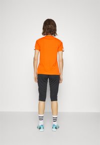Camisa polo deportiva naranja con detalles en blanco, combinada con pantalones negros de tres cuartos. El modelo lleva calcetines a rayas y zapatillas coloridas.
